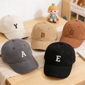 May include: Five baseball caps in various colors: beige, tan, brown, gray, and black. Each cap has a different embroidered letter: Y, B, G, A, and E. The caps are displayed on a wooden table.