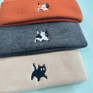 Puede incluir: Una pila de tres gorros de punto en naranja, gris y beige. Cada gorro presenta un dise&ntilde;o de gato pixelado bordado. Los gorros est&aacute;n hechos de un material de punto suave, perfecto para el clima fr&iacute;o.