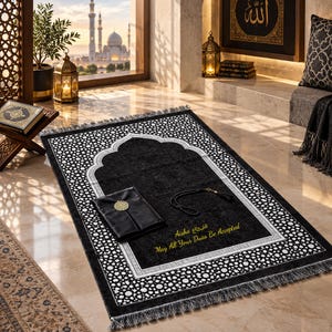 May include: A black prayer rug featuring a white geometric border, a central arch design, and gold embroidered text that reads Aisha and May All Your Duas Be Accepted. A black leather-bound book and black prayer beads rest on the rug surface.