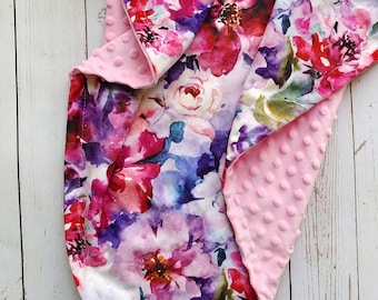 Puede incluir: Una manta de bebé rosa y blanca con flores y un respaldo suave y texturizado. La manta presenta un estampado floral de estilo acuarela con flores rosas, moradas y azules.