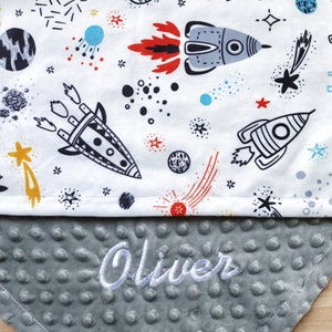 Op de afbeelding: Een grijze en witte babydeken met een ruimtethema print met raketten, sterren en planeten. De deken heeft een zachte, textuurrijke achterkant en is gepersonaliseerd met de naam "Oliver" geborduurd in wit.