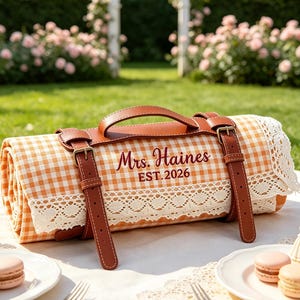 May include: A rolled picnic blanket featuring an orange and white gingham pattern with a white lace trim. The blanket is secured by brown leather straps and a handle, with the text Mrs. Haines EST. 2026 embroidered in dark brown on the front.