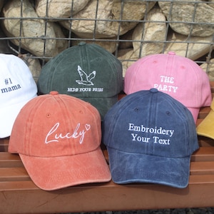 Puede incluir: Una colección de gorras de béisbol en varios colores, incluyendo blanco, verde, rosa, naranja, azul y amarillo. Cada gorra presenta texto bordado como "#1 mama", "Lucky" y "Embroidery Your Text". Las gorras se exhiben sobre una superficie de madera.