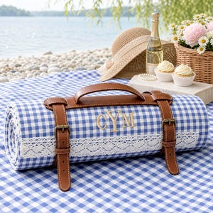 May include: A rolled blue and white gingham picnic blanket featuring a brown faux leather carrying handle and two buckled straps. The blanket is accented with a white lace trim and embroidered with the gold-coloured monogram letters CYM.