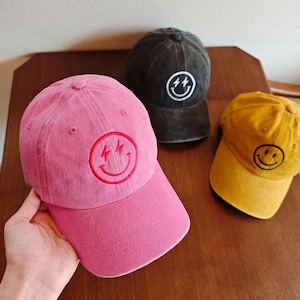 Könnte beinhalten: Drei Baseballkappen in Rosa, Schwarz und Gelb. Jede Kappe hat ein gesticktes Smiley-Gesicht mit Blitzaugen. Die Kappen sind aus einem weichen, strukturierten Stoff gefertigt und haben einen gebogenen Schirm.