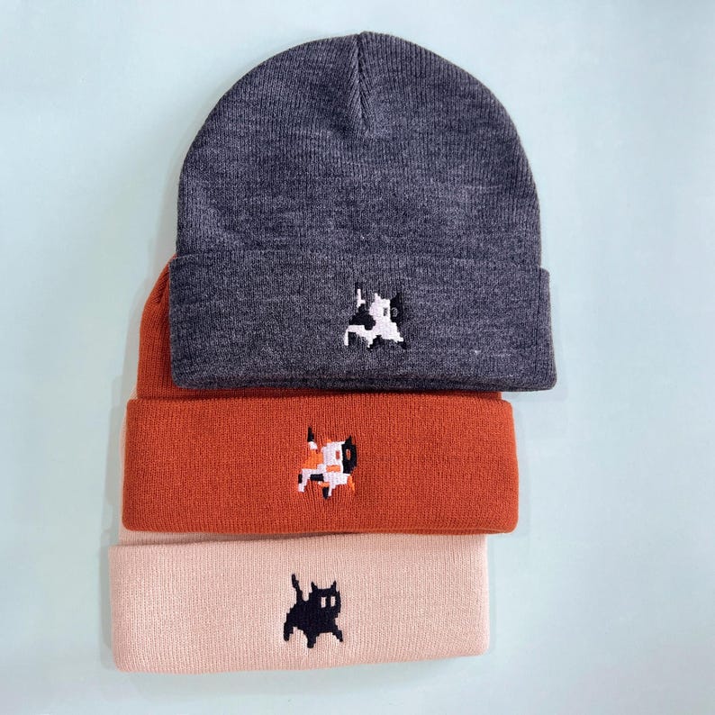 Puede incluir: Tres gorros de punto apilados. El gorro superior es gris oscuro con un dise&ntilde;o de perro pixelado blanco y negro. El gorro del medio es naranja con un dise&ntilde;o de perro pixelado. El gorro inferior es rosa claro con un dise&ntilde;o de gato pixelado negro.