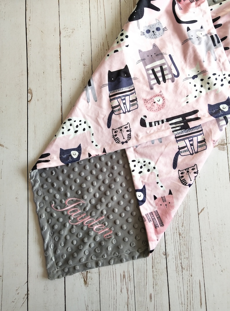 May include: A pink and gray baby blanket with a cat print on one side and a gray minky dot fabric on the other side. The blanket is personalized with the name "Jayden" embroidered on the gray side.