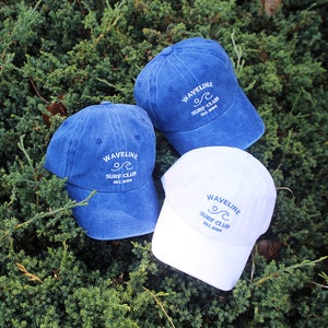 May include: Three denim baseball caps with white embroidered text that reads "Wavelline Surf Club Est. 2004". One cap is blue, one is white, and one is a darker blue.
