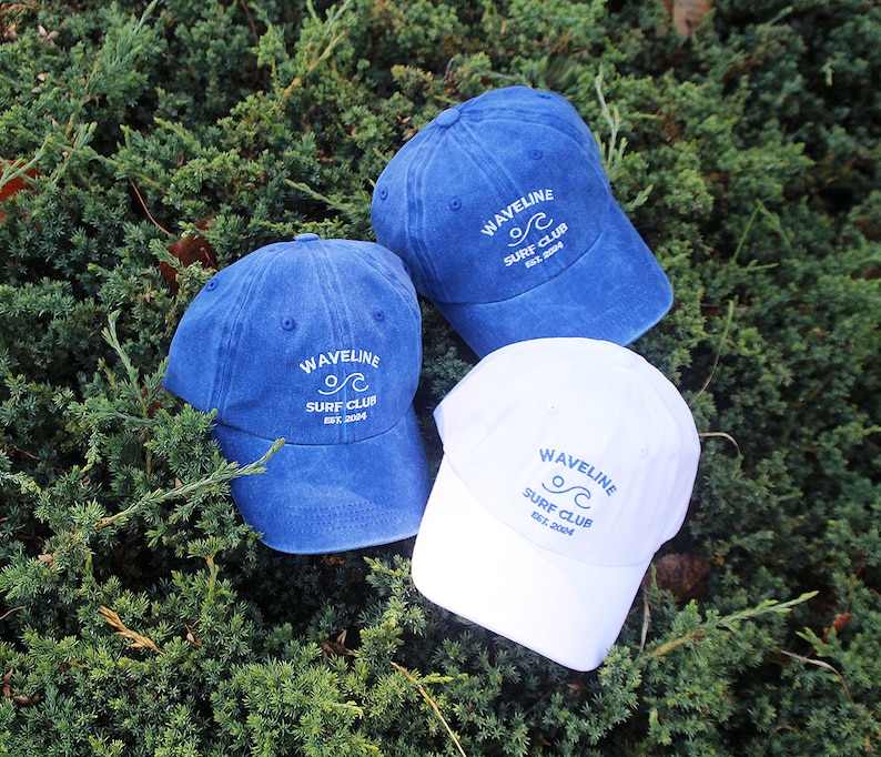 May include: Three denim baseball caps with white embroidered text that reads "Wavelline Surf Club Est. 2004". One cap is blue, one is white, and one is a darker blue.