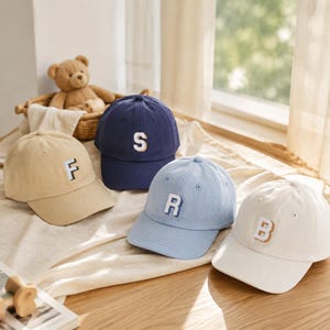 May include: Four baseball caps in beige, navy blue, light blue, and off-white. Each cap features a single, large, block-style letter embroidered on the front panel: F on the beige cap, S on the navy cap, R on the light blue cap, and B on the white cap.