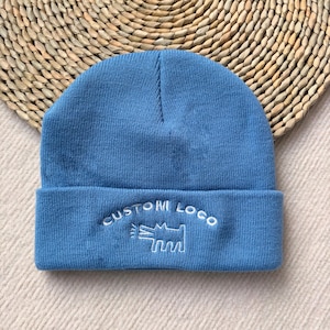 May include: A light blue beanie with a folded brim. The beanie features the embroidered text "CUSTOM LOGO" above a stylized dog illustration. The hat is made of a knitted material and is displayed on a woven surface.