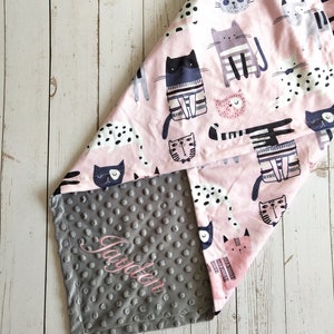 May include: A gray and pink baby blanket with a cat print on one side and a gray minky dot fabric on the other side. The gray side has the name "Jayden" embroidered on it.