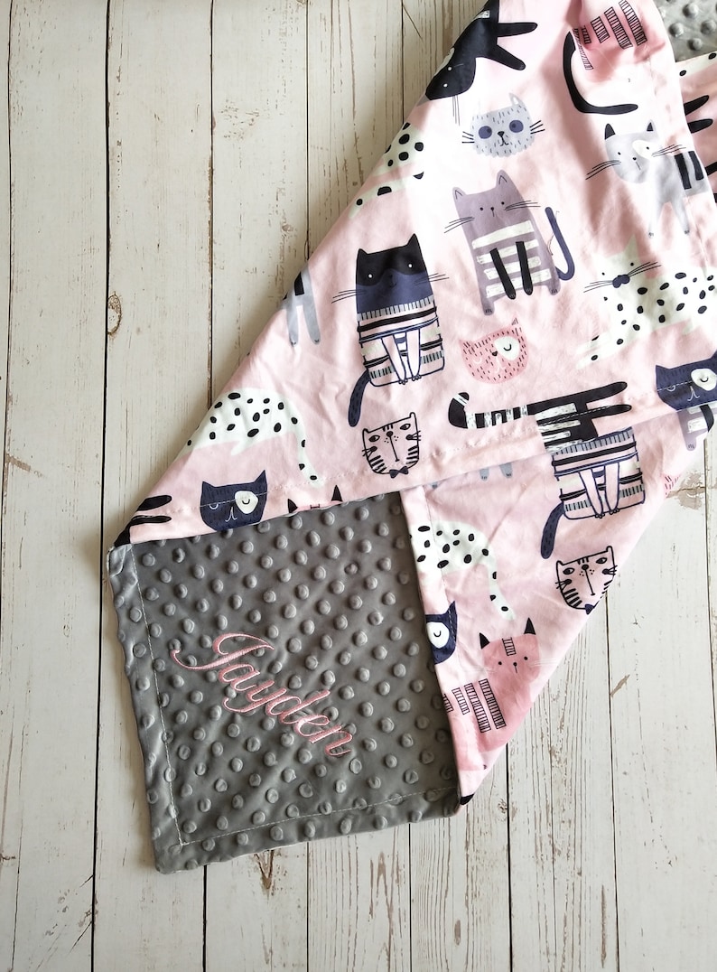May include: A gray and pink baby blanket with a cat print on one side and a gray minky dot fabric on the other side. The gray side has the name "Jayden" embroidered on it.