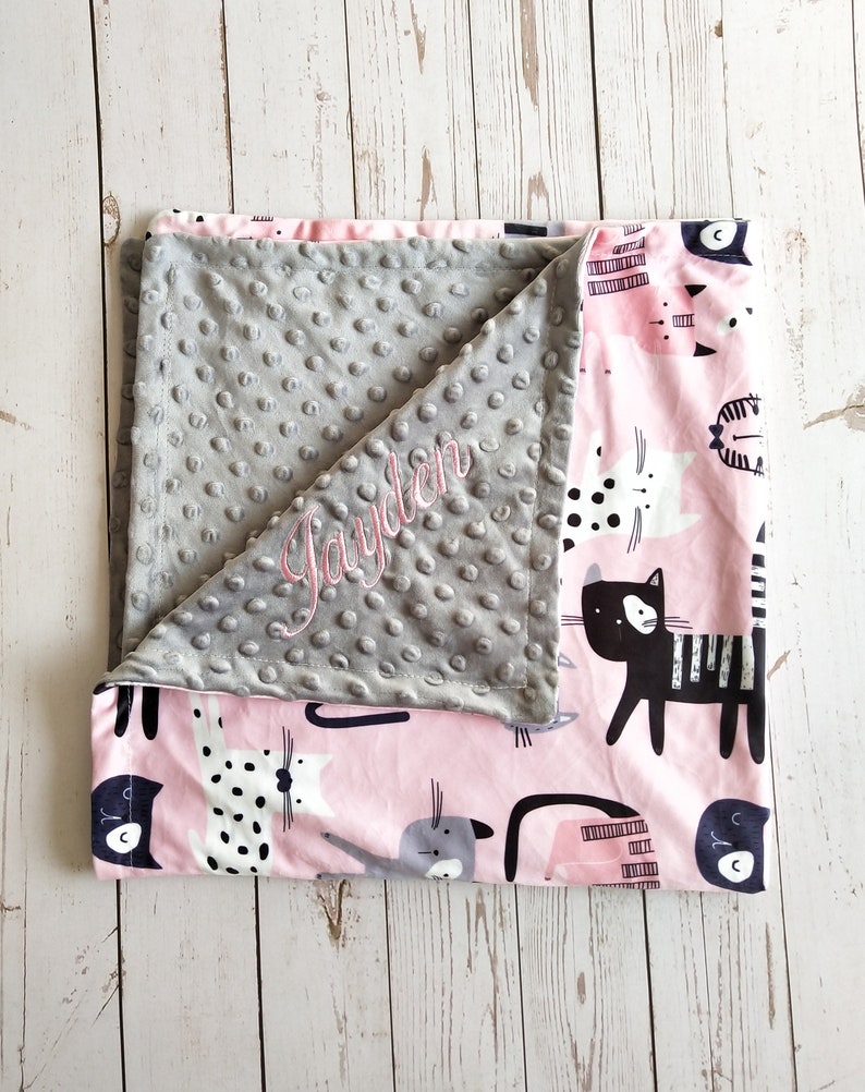 May include: A gray and pink baby blanket with a soft, textured side and a printed side featuring a pattern of black and white cats. The gray side is personalized with the name "Jayden" in pink embroidery.
