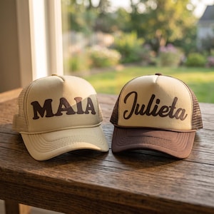 Puede incluir: Dos gorras tipo camionero con parte trasera de malla. La gorra de la izquierda es de color crema con el nombre MAIA en letras de bloque marrones. La de la derecha tiene un panel frontal crema con el nombre Julieta en cursiva marrón.