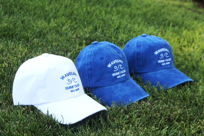 May include: Two baseball caps with the text "Wavelink Surf Club Est. 2004" embroidered on the front. One cap is white with blue embroidery and the other is blue with white embroidery.