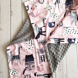 May include: A pink and gray reversible baby blanket with a cat print on one side. The cat print features various cats in different poses and patterns. The other side of the blanket is a soft gray with a textured pattern.