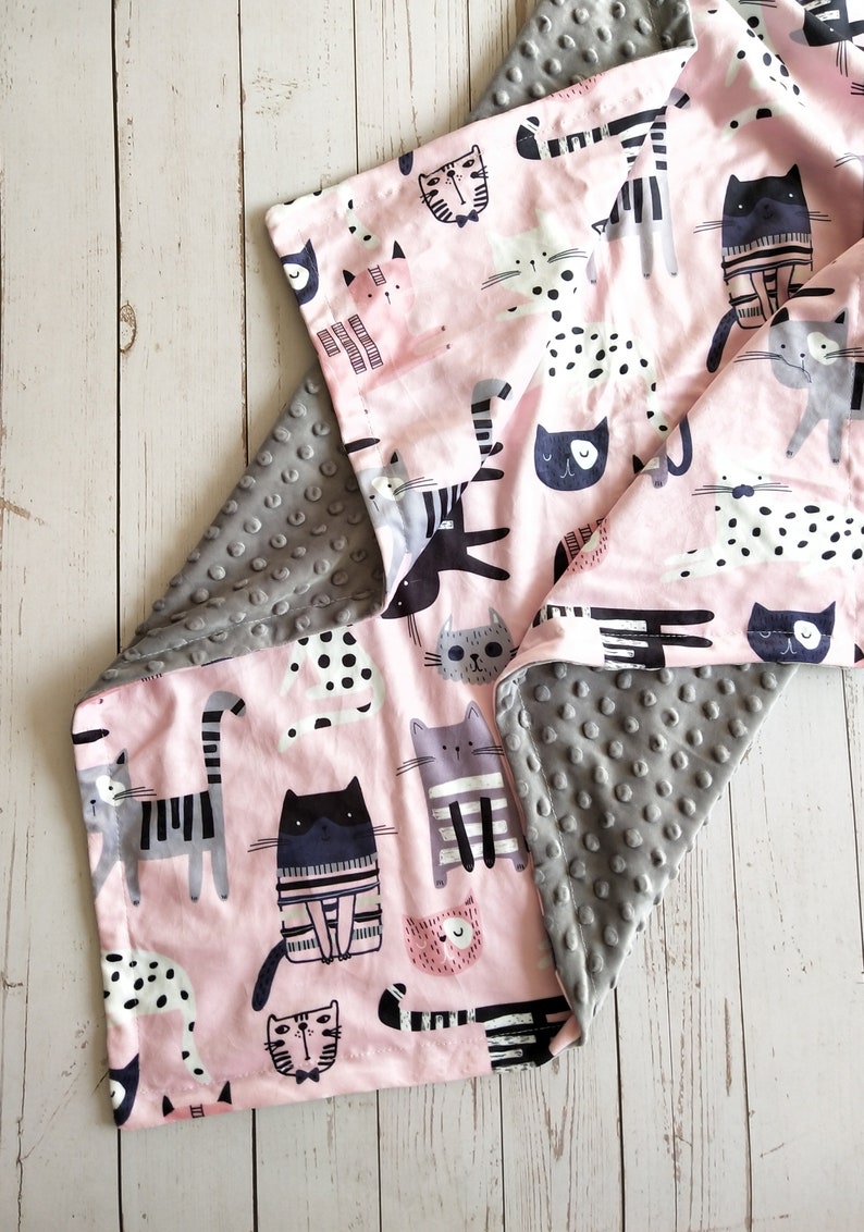 May include: A pink and gray reversible baby blanket with a cat print on one side. The cat print features various cats in different poses and patterns. The other side of the blanket is a soft gray with a textured pattern.