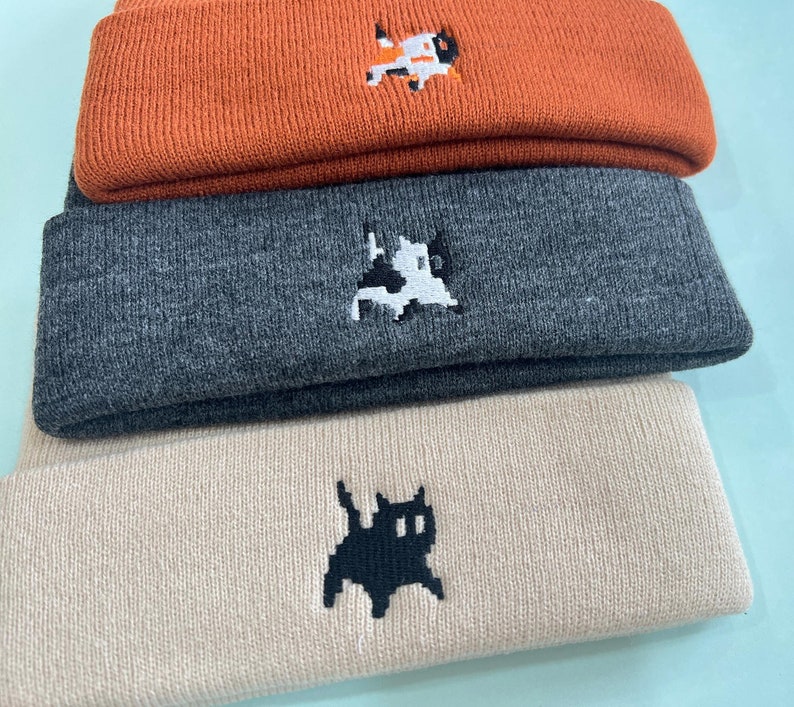 Puede incluir: Una pila de tres gorros de punto en naranja, gris y beige. Cada gorro presenta un dise&ntilde;o de gato pixelado bordado. Los gorros est&aacute;n hechos de un material de punto suave, perfecto para el clima fr&iacute;o.
