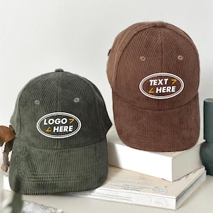 Puede incluir: Dos gorras de b&eacute;isbol de pana con dise&ntilde;os bordados diferentes. La gorra verde tiene "LOGO HERE" en un &oacute;valo blanco con detalles amarillos. La gorra marr&oacute;n tiene "TEXT HERE" en un &oacute;valo blanco con detalles amarillos.