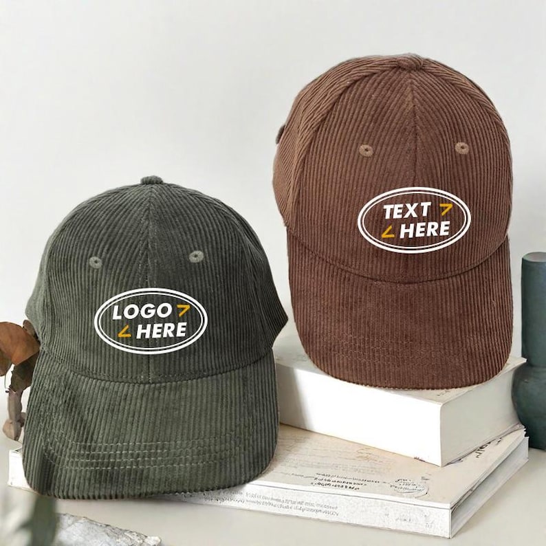 Puede incluir: Dos gorras de b&eacute;isbol de pana con dise&ntilde;os bordados diferentes. La gorra verde tiene "LOGO HERE" en un &oacute;valo blanco con detalles amarillos. La gorra marr&oacute;n tiene "TEXT HERE" en un &oacute;valo blanco con detalles amarillos.