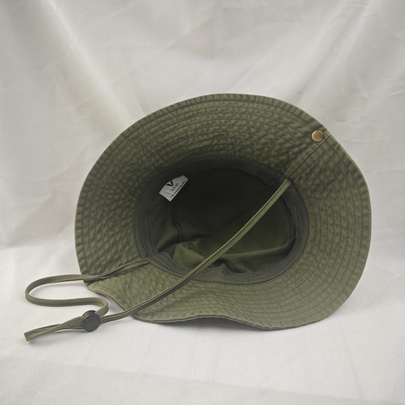 K&ouml;nnte beinhalten: Ein gr&uuml;ner Bucket-Hut mit einem braunen Band und einem schwarzen Knopf. Der Hut hat ein kleines Etikett innen, auf dem "VINTAGE" steht.