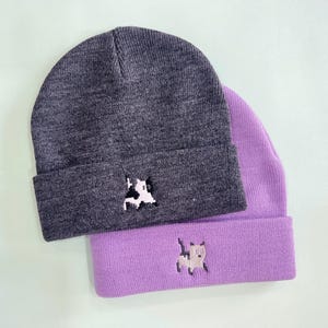 Puede incluir: Dos gorros de punto, uno gris carb&oacute;n y otro lavanda, est&aacute;n apilados. Cada gorro presenta un peque&ntilde;o dise&ntilde;o de vaca pixelada bordado en el borde doblado. Los gorros est&aacute;n hechos de un material de punto suave.
