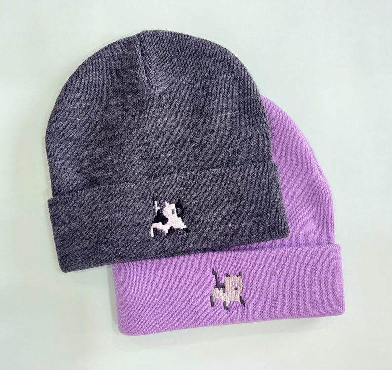 Puede incluir: Dos gorros de punto, uno gris carb&oacute;n y otro lavanda, est&aacute;n apilados. Cada gorro presenta un peque&ntilde;o dise&ntilde;o de vaca pixelada bordado en el borde doblado. Los gorros est&aacute;n hechos de un material de punto suave.