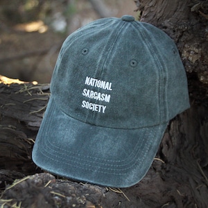 May include: A dark green baseball cap with the text "NATIONAL SARCASM SOCIETY" embroidered in white on the front.