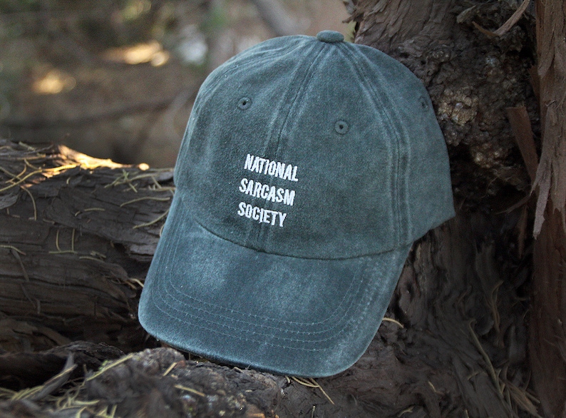 May include: A dark green baseball cap with the text "NATIONAL SARCASM SOCIETY" embroidered in white on the front.