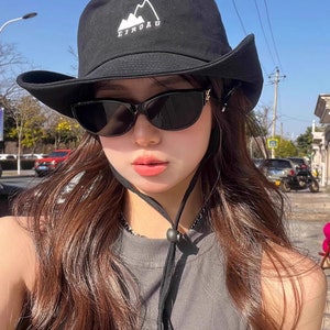 May include: A black bucket hat with a white mountain logo and the text "EIROAU" embroidered on the front. The hat has a wide brim and a black chin strap. The person wearing the hat is also wearing black sunglasses.