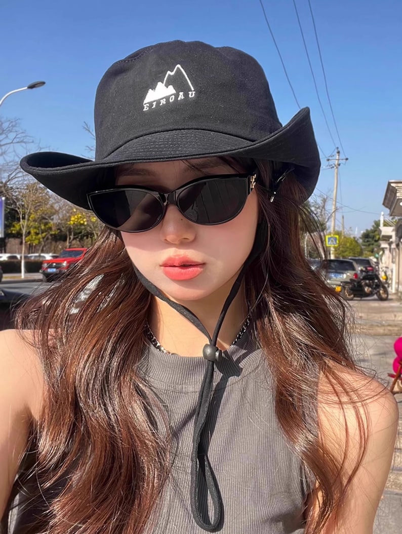 May include: A black bucket hat with a white mountain logo and the text "EIROAU" embroidered on the front. The hat has a wide brim and a black chin strap. The person wearing the hat is also wearing black sunglasses.