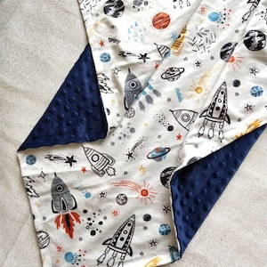 May include: A white baby blanket with a navy blue minky backing. The blanket features a space-themed print with rockets, stars, planets, and comets.