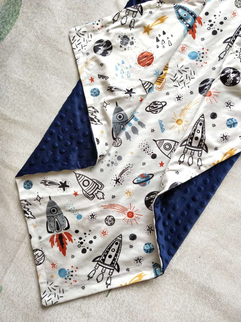 May include: A white baby blanket with a navy blue minky backing. The blanket features a space-themed print with rockets, stars, planets, and comets.