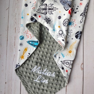 May include: A gray and white baby blanket with a space theme. The blanket has a soft, textured side and a printed side with rockets, stars, and planets. The blanket is personalized with the name "Matias Lim" in white embroidery.