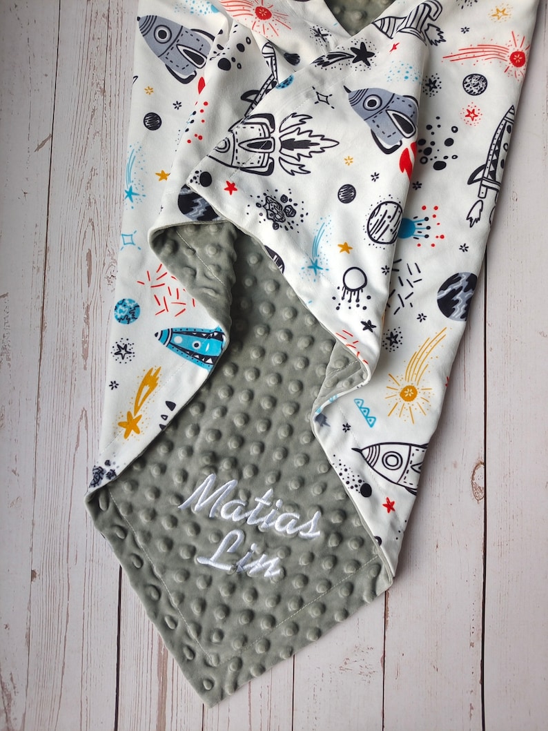 May include: A gray and white baby blanket with a space theme. The blanket has a soft, textured side and a printed side with rockets, stars, and planets. The blanket is personalized with the name "Matias Lim" in white embroidery.