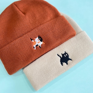 Puede incluir: Dos gorros de punto, uno naranja quemado y otro beige, est&aacute;n apilados. Cada gorro presenta un dise&ntilde;o de gato bordado. El gorro naranja tiene un gato calic&oacute;, mientras que el gorro beige tiene un gato negro.