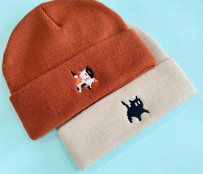 Puede incluir: Dos gorros de punto, uno naranja quemado y otro beige, est&aacute;n apilados. Cada gorro presenta un dise&ntilde;o de gato bordado. El gorro naranja tiene un gato calic&oacute;, mientras que el gorro beige tiene un gato negro.