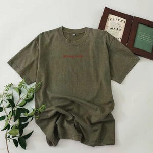 Puede incluir: Camiseta verde oliva de manga corta. El texto "Healing is hot" está impreso en rojo en el pecho. La camiseta tiene un aspecto ligeramente arrugado, lo que sugiere un estilo informal. Una imagen enmarcada está en el fondo.