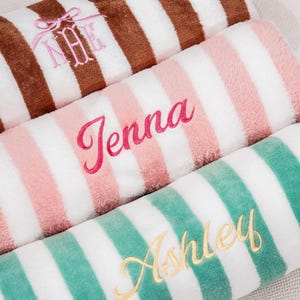 Könnte beinhalten: Nahaufnahme von drei gerollten, gestreiften Strandtüchern. Das obere Handtuch ist braun und weiß mit gestickten Initialen. Das mittlere Handtuch ist rosa und weiß mit dem Namen "Jenna". Das untere Handtuch ist grün und weiß mit dem Namen "Ashley".