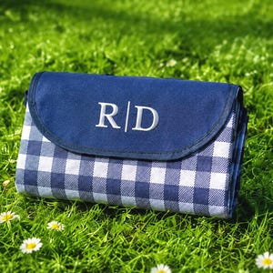 Può includere: Una coperta da picnic rettangolare piegata con un motivo a quadretti vichy blu navy e bianco. La patta superiore è blu navy tinta unita e presenta le lettere bianche ricamate R | D centrate sulla parte anteriore, arrotolata in modo compatto.