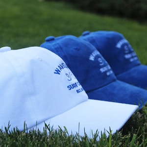 May include: Three baseball caps, one white and two blue, with the text "Wavecrest Surf Co. Est. 1972" embroidered on the front.