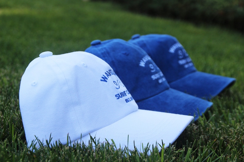 May include: Three baseball caps, one white and two blue, with the text "Wavecrest Surf Co. Est. 1972" embroidered on the front.