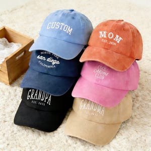 May include: A stack of six baseball caps in various colors, including blue, orange, pink, navy, black, and tan. Each cap features white embroidered text, such as "CUSTOM," "MOM," "COOL Moms CLUB," "GRANDPA," "GRANDMA," and "NORTH PARK san diego CALIFORNIA."