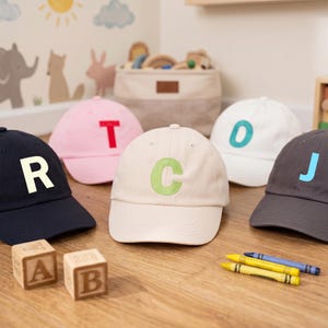 May include: A collection of five baseball caps in navy, pink, cream, white, and charcoal gray. Each cap features a single large, bold letter on the front: R in cream, T in red, C in light green, O in teal, and J in light blue, all in a classic style.