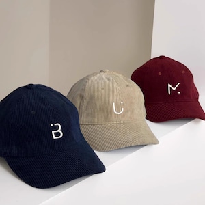 Puede incluir: Tres gorras de b&eacute;isbol de pana en diferentes colores: azul marino, beige y burdeos. Cada gorra tiene una letra bordada en blanco en la parte delantera: "B", "U" y "M".