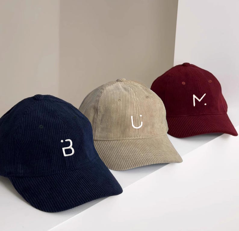 Puede incluir: Tres gorras de b&eacute;isbol de pana en diferentes colores: azul marino, beige y burdeos. Cada gorra tiene una letra bordada en blanco en la parte delantera: "B", "U" y "M".