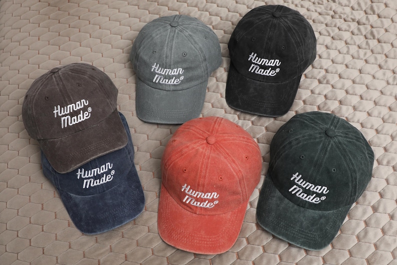 May include: Six baseball caps in various colors, including black, gray, blue, and red. Each cap features the embroidered text "Human Made" in white.