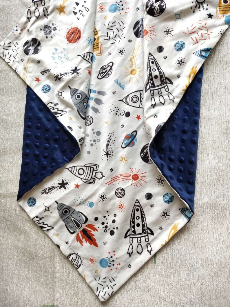 May include: A white baby blanket with a navy blue backing. The blanket features a space-themed print with rockets, stars, planets, and comets.