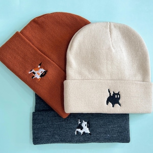 Puede incluir: Tres gorros de punto en &oacute;xido, crema y gris carb&oacute;n. Cada gorro presenta un peque&ntilde;o dise&ntilde;o bordado: un gato, una vaca y una figura geom&eacute;trica. Los gorros est&aacute;n doblados en el borde.